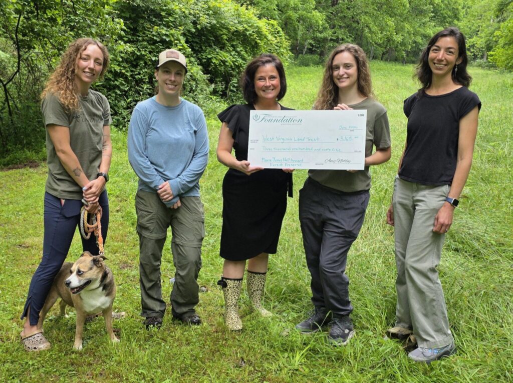 Group of six people outdoors in a grassy, wooded area holding a large donation check; a brown dog sits at the front left.