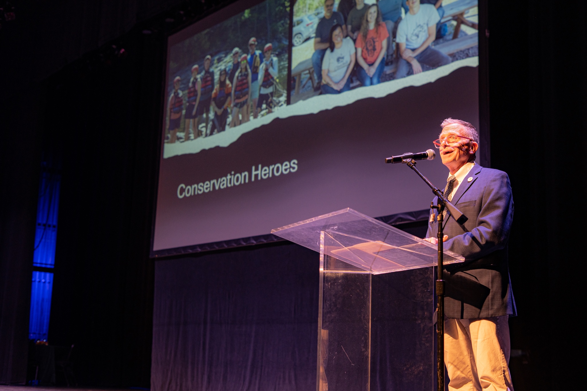 Man in a suit speaking at a clear podium on stage, large screen behind showing a collage and the words 'Conservation Heroes'